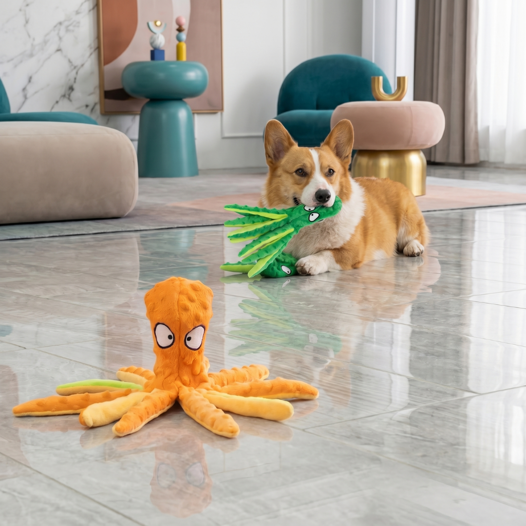 Orange plush octopus dog toy with a corgi chewing a green squeaky toy in the background.
