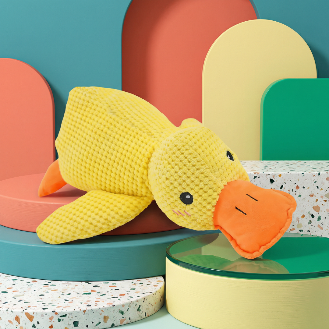 Yellow plush duck dog toy with an orange beak and feet, resting on colorful geometric blocks.