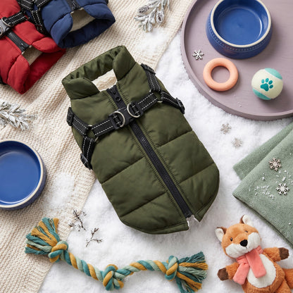 Olive green puffer dog coat with built-in harness, surrounded by pet bowls, toys, and winter decor.